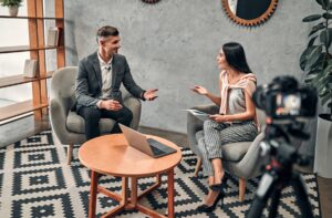 An interviewer speaking with a business guest while a camera records